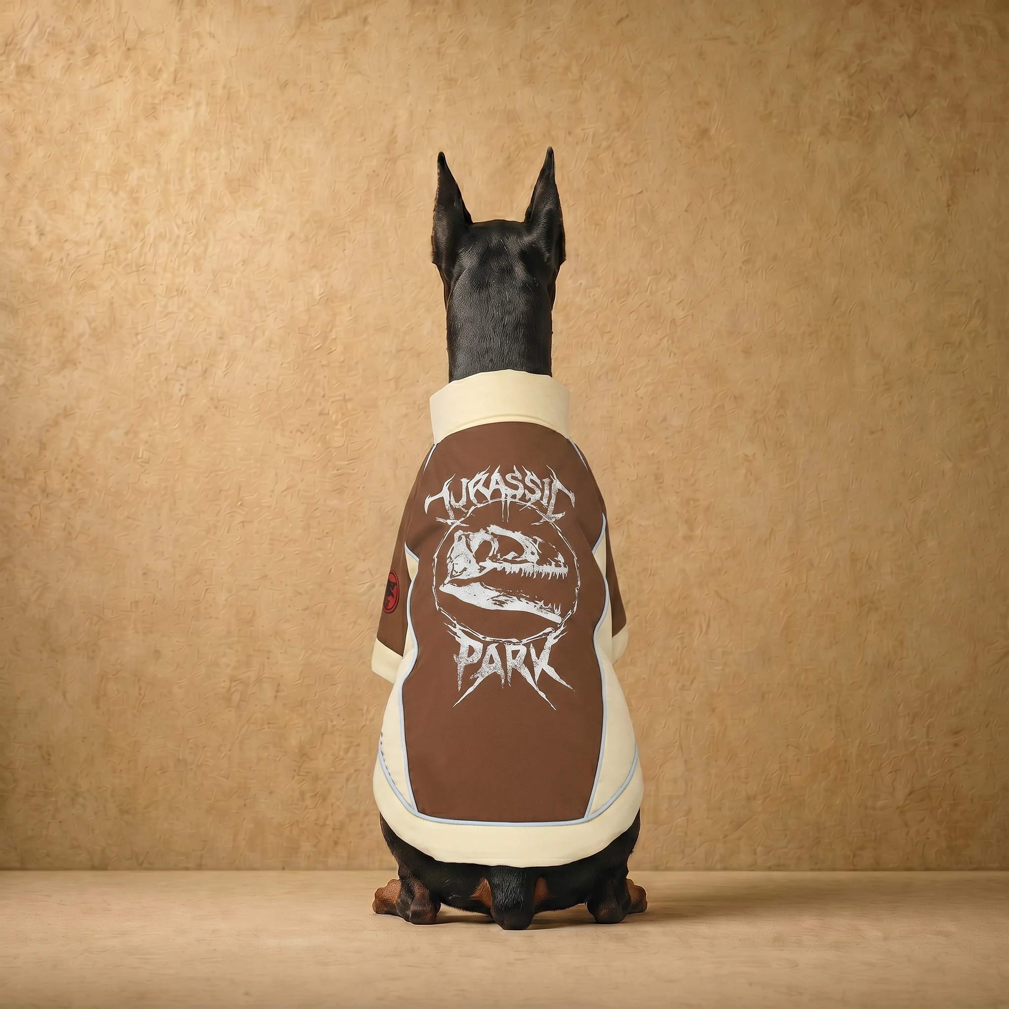 Doberman wearing a brown and cream insulated coat with a white dinosaur skull graphic, designed for deep-chested dogs, shown from behind against a textured beige backdrop.